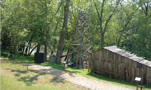 Heavily wooded Burning Springs, W.V., park along creek with small, wooden derrick and displays.