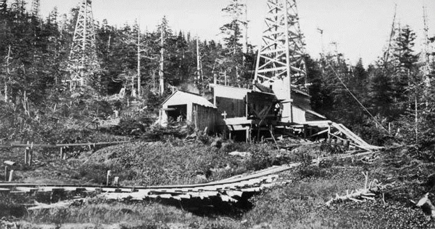 Photo of a small Alaska railroad leading to oil derricks in mountains.