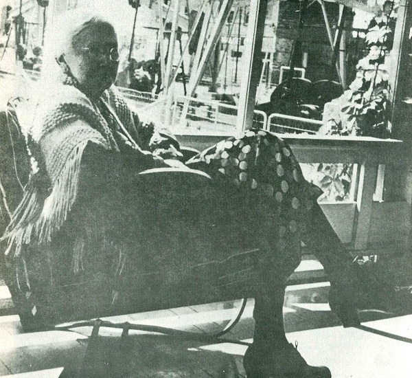 Mrs. Lou Della Crim sitting and wearing a shawl, watches her nearby pumping oil wells from her porch.