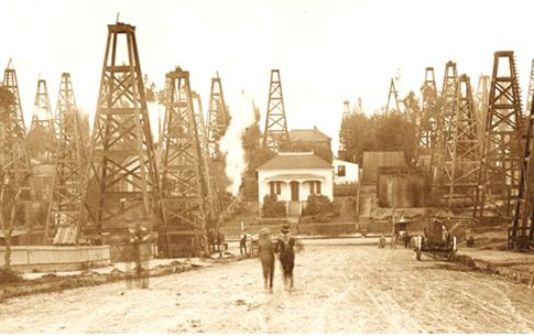 Newsletter-Oil-Queen-LA-AOGHS A sepia image of two men standing with linked arms among wooden derricks on a road leading to a house in the Los Angeles City oilfield at the turn of the century.